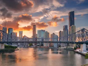 A panoramic view of the Brisbane River with the iconic Story Bridge arching over it. The modern city skyline is visible in the background, reflecting on the calm waters. The foreground features lush greenery from the riverside parks. The sky is adorned with a warm sunset glow, casting golden hues over the scene.