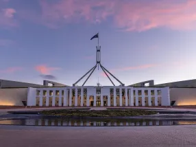 The modern Parliament House situated on Capital Hill, featuring its distinctive flag mast. The building is surrounded by impeccably maintained gardens and pathways. In the foreground is Lake Burley Griffin with kayaks and sailboats on the water. The Australian War Memorial can be subtly included in the background. The sky is clear, highlighting a crisp day.