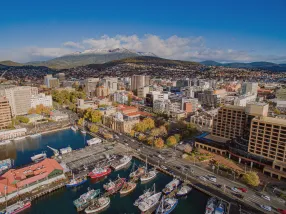 The charming Salamanca Place with its row of Georgian sandstone warehouses converted into cafes and art galleries. The backdrop features Mount Wellington, its peak possibly dusted with snow. The nearby harbor has classic sailing boats and fishing vessels. The atmosphere is cozy with a touch of historic charm.