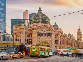 A bustling city street showcasing Melbourne's famous trams traveling down the road. On one side is the historic Flinders Street Station with its ornate architecture; on the other, modern skyscrapers. Vibrant street art adorns nearby laneways, adding splashes of color. People are sitting at outdoor cafes, emphasizing the city's lively atmosphere.