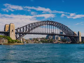 The majestic Sydney Opera House with its distinctive white sails, set against the backdrop of the Sydney Harbour Bridge. The shimmering harbor waters have ferries and sailboats gliding across. The city skyline rises in the background. The sky is a vibrant blue with a few fluffy clouds, emphasizing a bright and sunny day.